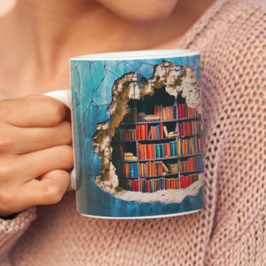 Op de afbeelding: Witte keramische mok met een ontwerp van een boekenkast zichtbaar door een gat in een blauwe muur. De boekenkast is gevuld met kleurrijke boeken. De mok wordt vastgehouden door een persoon die een roze trui draagt.