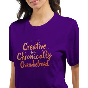 Op de afbeelding: Een paars T-shirt met de gouden tekst "Creative but Chronically Overwhelmed". De tekst is in een handschriftstijl. Het T-shirt heeft een ronde hals en korte mouwen. De tekst is gecentreerd op de borst.