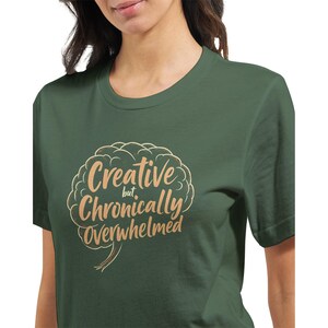 May include: Green t-shirt with the phrase "Creative but Chronically Overwhelmed" in a light brown, brain-shaped design. The text is in a handwritten style. The shirt is made of a soft material.
