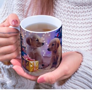 Op de afbeelding: Witte keramische mok met een kerstthema met twee golden retriever puppy's, een rode strik en een gouden geschenkdoos. De mok is gevuld met een lichtbruine vloeistof.