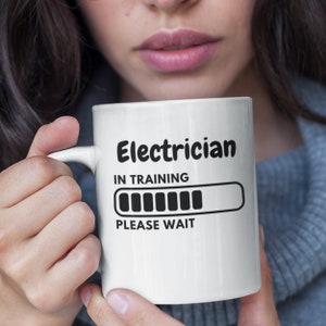 Peut inclure: Une tasse en céramique blanche avec un motif noir qui dit "Electricien en formation" avec une barre de progression presque pleine et le texte "Veuillez patienter".