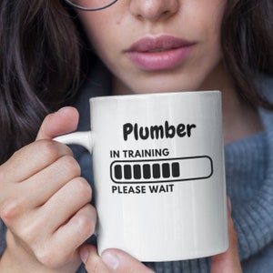 Puede incluir: Taza de cerámica blanca con el texto "Plumber IN TRAINING PLEASE WAIT" en negro. La taza presenta un gráfico de barra de carga. La taza es sostenida por una persona que lleva un suéter gris.