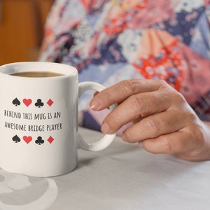May include: White ceramic mug with black text that reads "Behind this mug is an awesome bridge player" and four playing card symbols: a spade, heart, club, and diamond.