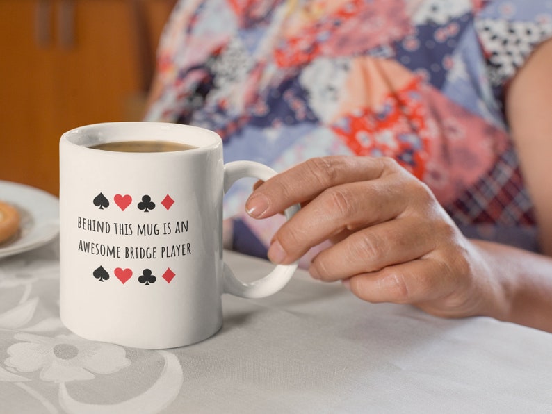 May include: White ceramic mug with black text that reads "Behind this mug is an awesome bridge player" and four playing card symbols: a spade, heart, club, and diamond.