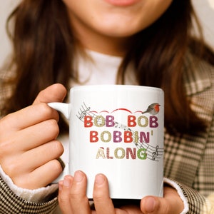 Poczuj świąteczną atmosferę z naszym świątecznym kubkiem: „Bob Bob Bobbin' Along” z wielokolorowymi czcionkami w stylu retro, uroczym rudzikiem i muzycznymi nutami!