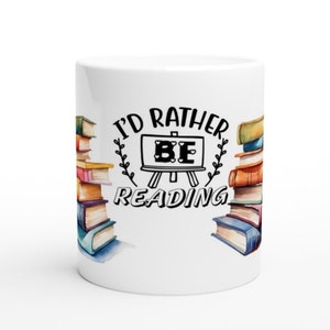 Puede incluir: Taza de cerámica blanca con un gráfico en blanco y negro que dice "I'd Rather Be Reading" con un dibujo en blanco y negro de un caballete con la palabra "BE" en él. La taza está decorada con una ilustración de acuarela de una pila de libros coloridos a cada lado.
