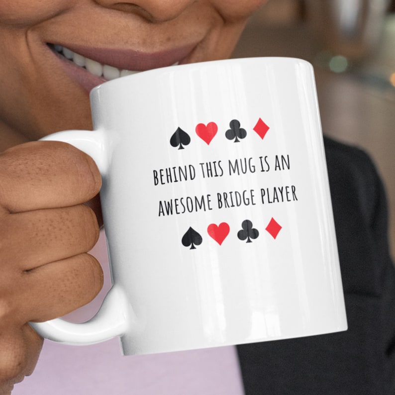 May include: White ceramic mug with black text that reads "Behind this mug is an awesome bridge player". The text is surrounded by four black playing card suits and four red playing card suits.