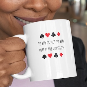 May include: White ceramic coffee mug with a black and red design of playing card suits and the text "To bid or not to bid that is the question".