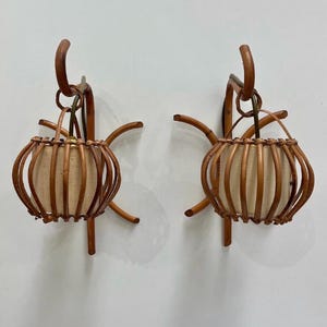 May include: Two matching wall sconces with a woven rattan design. Each sconce features a cylindrical, off-white fabric shade encased in a brown rattan cage. The sconces have curved, brown metal arms for mounting.