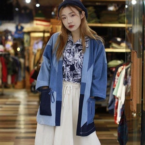 Op de afbeelding: Een vrouw draagt een blauw denim jack met een patchwork ontwerp. Het jack wordt gedragen over een wit shirt met bloemenprint en een witte rok. Ze draagt een blauwe beanie.