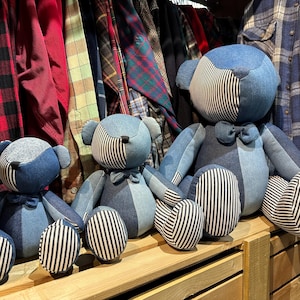 Può includere: Tre orsacchiotti di denim di diverse dimensioni, seduti su uno scaffale di legno. Gli orsi sono fatti di diverse tonalità di denim blu e hanno strisce bianche.