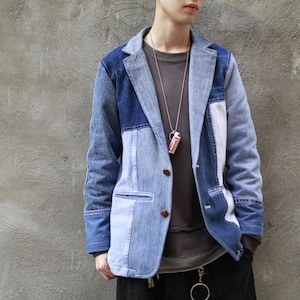 May include: A blue denim jacket with a patchwork design. The jacket has a button closure and two pockets. The jacket is worn over a grey t-shirt.