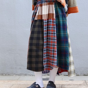 May include: A patchwork skirt with panels of different plaid patterns in green, red, brown, and navy blue. The skirt has a button-down front and an elasticated waistband. The person is wearing white socks and denim clogs.