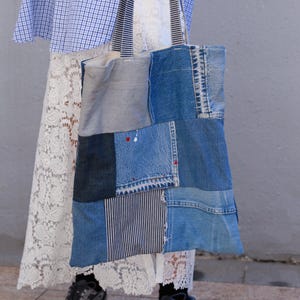 May include: A patchwork tote bag made from various denim fabrics in shades of blue, gray, and white. The bag features a mix of textures and patterns, including stripes and solid denim. The handles are striped, and the bag is held by a person wearing a white lace skirt.