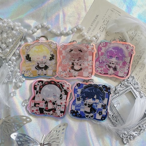 May include: Five colorful keychains featuring anime characters with different names and designs. Each keychain has a white background with black and white checkered patterns and pink flowers. The characters are dressed in maid outfits with different colored dresses and hair.