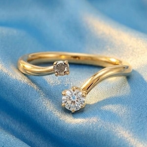 May include: A gold-colored ring with two gemstones. One gemstone is a small, dark gray square, and the other is a larger, round, clear gemstone. The ring is set against a blue satin background.