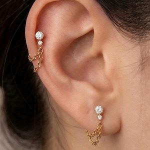 May include: A pair of gold-toned earrings featuring two clear, round-cut stones of varying sizes set in a vertical arrangement, connected by a delicate gold chain that loops back to the post, creating a draped, linked aesthetic on the ear.