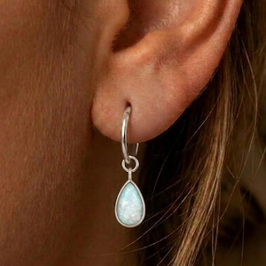 Può includere: Un orecchino a cerchio in argento con una pietra preziosa opale a forma di goccia pendente. L'orecchino è fissato al lobo dell'orecchio e mette in risalto i riflessi iridescenti bianchi e blu sulla superficie liscia e lucida.