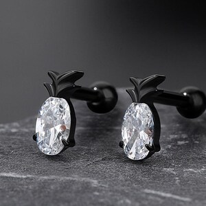 May include: A pair of black barbell earrings. Each earring features a black pineapple design with a clear, oval-cut stone. The earrings are displayed on a gray, textured surface.