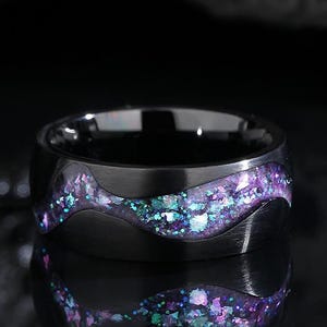 Puede incluir: Un anillo negro con una incrustación en forma de ola de purpurina iridiscente púrpura, azul y verde. El anillo tiene un acabado de metal cepillado y una banda ancha. El anillo se muestra sobre una superficie reflectante.