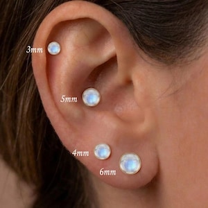 Peut inclure: Quatre boucles d'oreilles à clous en pierre de lune ronde serties dans du métal doré, présentées sur une oreille. Les clous sont étiquetés avec les tailles 3mm, 4mm, 5mm et 6mm. Chaque boucle présente une pierre de lune lisse et irisée.