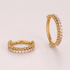 May include: Two gold-toned hoop rings featuring a dual-row design. One row consists of small, polished gold spheres, while the adjacent row is set with a continuous line of clear, sparkling round-cut stones. The hoops have a hinged closure.