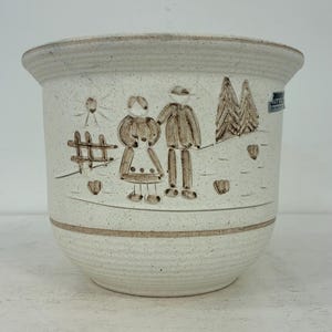 Op de afbeelding: Een gespikkelde, gebroken witte keramieken bloempot met een bruine, handgeschilderde illustratie van twee figuren bij een hek, een zon en twee bomen. De pot heeft een brede, uitlopende rand, een ronde vorm en een horizontale bruine lijn.