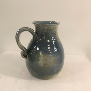 Op de afbeelding: Een keramische kan met een rond lichaam en een gebogen handvat met een decoratieve krul aan de onderkant. Het oppervlak heeft een glanzend glazuur in tinten diepblauw, leigrijs en gedempt groen, met zichtbare horizontale ribbels langs de hals.