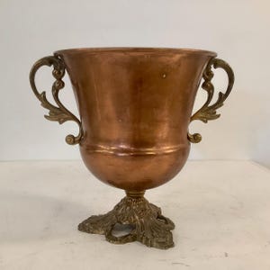 Puede incluir: Una urna de color cobre con un borde ancho y acampanado y un cuerpo liso y redondeado. El recipiente cuenta con dos asas ornamentadas en forma de voluta a cada lado y descansa sobre una base decorativa tipo pedestal con detalles de hojas.