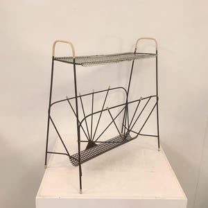 Puede incluir: Un revistero de metal negro con un estante superior que presenta una superficie de malla blanca y dos asas arqueadas de color claro. La sección inferior tiene una base de rejilla de metal negro curvada con soportes de alambre.