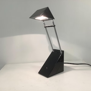 May include: A black desk lamp featuring a triangular base and a matching triangular head. The lamp has two thin, parallel silver metal arms connecting the base to the head, which emits a bright light from its underside. A black power cord is attached.