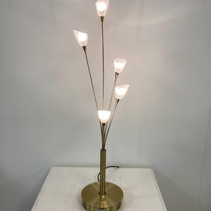 Pode incluir: Um candeeiro de pé em latão com uma base circular e um poste central que se ramifica em cinco braços finos. Cada braço termina num pequeno abajur de vidro branco em forma de tulipa, contendo cada um uma lâmpada acesa.