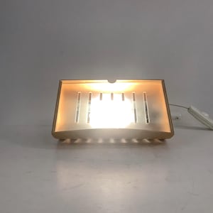 May include: A rectangular wall-mounted lamp with a light-coloured frame and a translucent front panel featuring vertical cutouts. The lamp emits a warm, bright glow from within and includes a white electrical cord with an attached power switch.