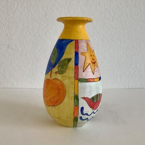 Pode incluir: Um vaso de cerâmica com pescoço amarelo e corpo dividido em painéis com ilustrações pintadas à mão. Os desenhos incluem uma fruta laranja com folhas verdes, um sol amarelo com um rosto e um pássaro vermelho sobre linhas.