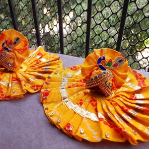 Puede incluir: Dos tocados de pavo real de tela amarilla y dorada con detalles de joyas. Los tocados están decorados con patrones blancos y rojos.