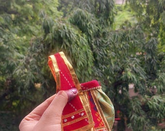 May include: A small, ornate red and gold fabric item with a decorative design. The item has gold trim and small, clear embellishments. The background is a blurred view of green foliage.