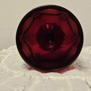 Op de afbeelding: Een robijnrode glazen beker met een ruitpatroon rond de rand. De beker staat op een witte, geborduurde doek. Het glas is transparant en laat licht door.