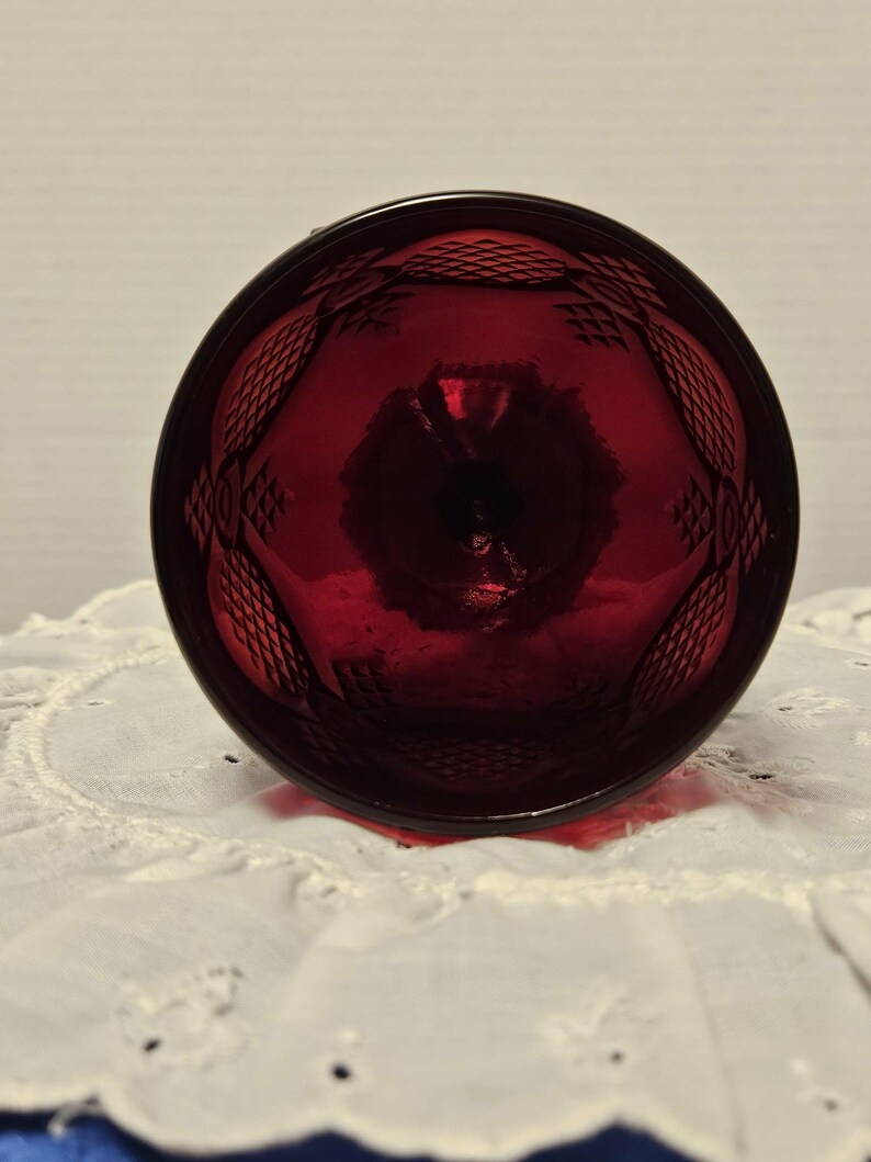 Op de afbeelding: Een robijnrode glazen beker met een ruitpatroon rond de rand. De beker staat op een witte, geborduurde doek. Het glas is transparant en laat licht door.