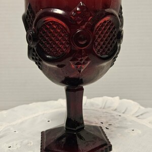 Op de afbeelding: Een robijnrode vintage kelk met een gedetailleerd patroon. Het glas heeft een diamant- en cirkelontwerp op de kom, een steel en een vierkante voet. De kelk is waarschijnlijk gemaakt van persglas en is geschikt voor het serveren van dranken.