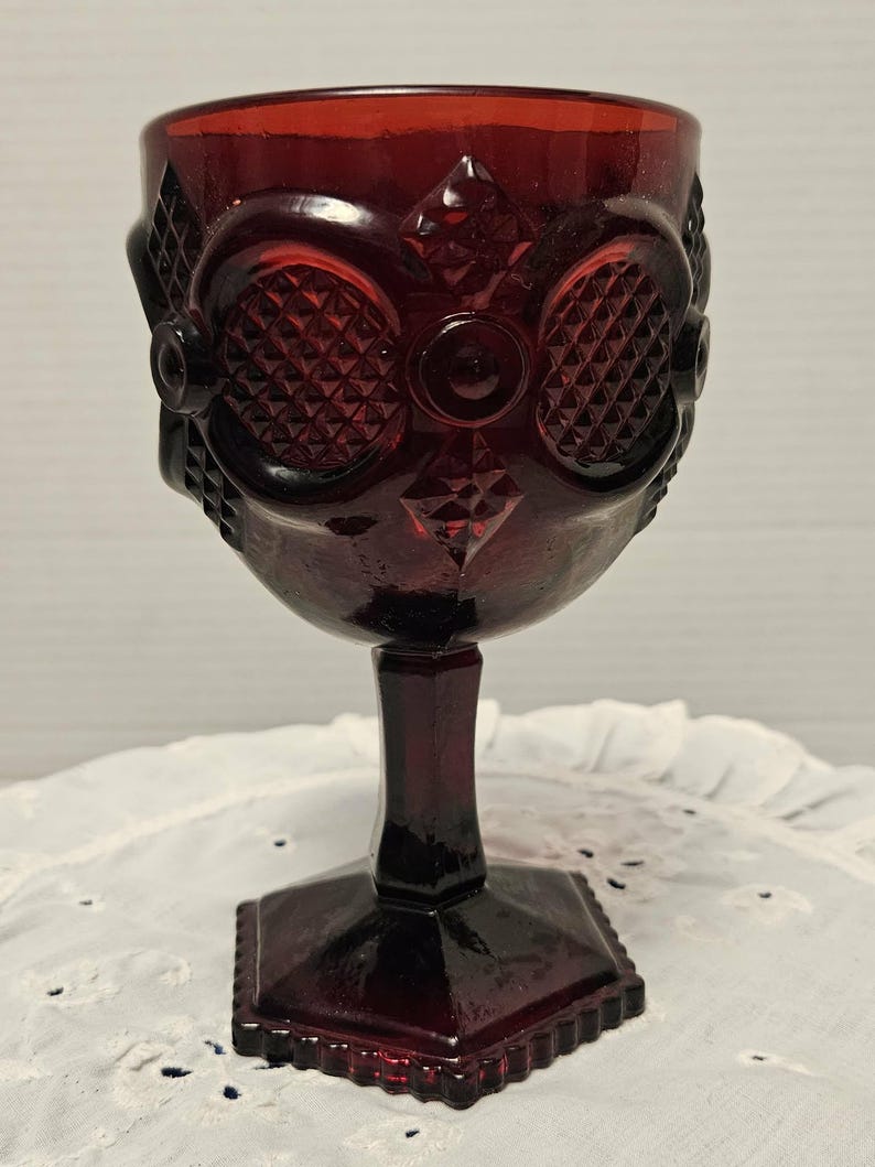 Op de afbeelding: Een robijnrode vintage kelk met een gedetailleerd patroon. Het glas heeft een diamant- en cirkelontwerp op de kom, een steel en een vierkante voet. De kelk is waarschijnlijk gemaakt van persglas en is geschikt voor het serveren van dranken.