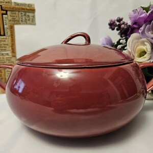 May include: A burgundy ceramic soup tureen with a matching lid and two side handles. The tureen has a glossy finish and a small loop handle on the lid. A decorative cross and flowers are in the background.