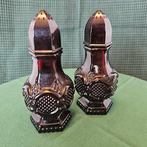 May include: A pair of dark purple glass salt and pepper shakers. Each shaker has a faceted, cone-shaped top with small holes. The bodies feature a textured pattern and sit on a hexagonal base.