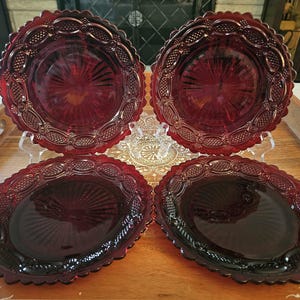 May include: A set of four deep red glass plates featuring a pressed pattern with a scalloped edge and a central sunburst design. The plates have a textured border with repeating oval and diamond motifs, creating a decorative rim around the center.