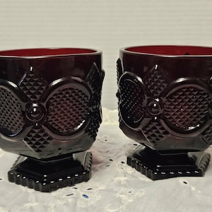 May include: Two ruby red glass goblets with intricate diamond and oval patterns. The goblets have a square base and a detailed design around the bowl. The glass has a glossy finish.