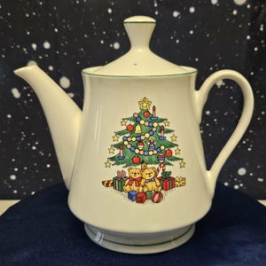 May include: White ceramic teapot with a Christmas tree design. The teapot features a green trim and a lid with a small handle. The Christmas tree is decorated with ornaments, and there are two teddy bears and wrapped gifts at the base.