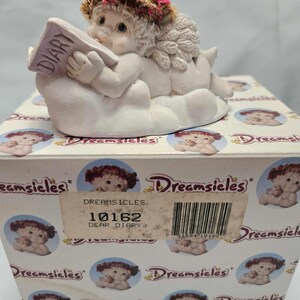 May include: A white porcelain figurine of an angel with a pink flower crown, reading a purple book titled "Diary" while lying on a white cloud. The figurine is on a white box with the text "Dreamsicles" and the product number "10162" printed on it.