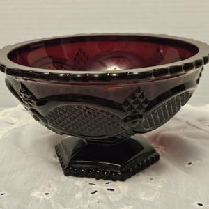 May include: A ruby red glass pedestal bowl with a decorative rim and ornate detailing. The bowl features a diamond pattern and floral accents, resting on a square base. This vintage piece is ideal for serving or display.