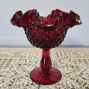 May include: A deep red glass compote dish featuring a hobnail texture on the exterior bowl. The rim is ruffled and crimped with a smooth finish. The piece stands on a circular pedestal base and a short, thick stem, all in a matching translucent red.