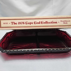 May include: A ruby red glass condiment dish from the Avon 1876 Cape Cod Collection. The dish is divided into two sections and has a decorative scalloped edge. The box above the dish reads "Avon The 1876 Cape Cod Collection Condiment Dish."