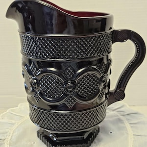 May include: A dark ruby-colored glass pitcher with a textured, diamond-patterned design. The pitcher features a spout, a handle, and decorative floral and geometric patterns. The base is a square shape.
